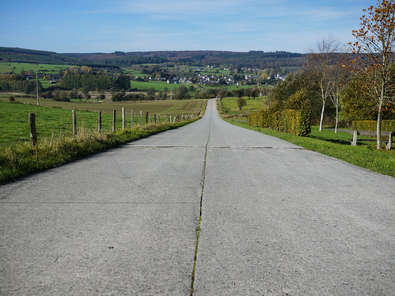 A long, straight concrete road stretches downhill toward a small village surrounded by green fields and trees.