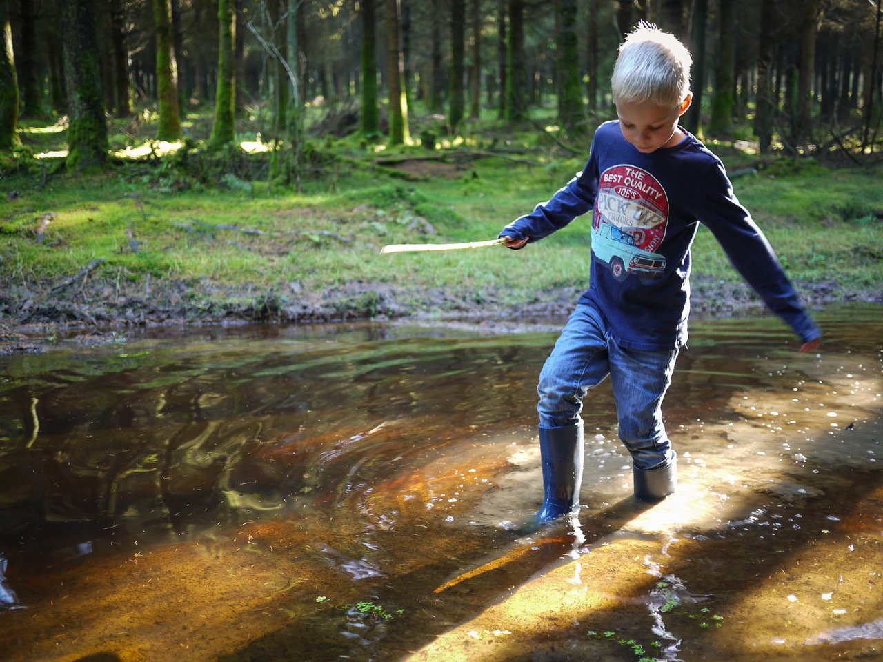 A young boy in rubber boots walks through a shallow forest stream, holding a stick in one hand.
