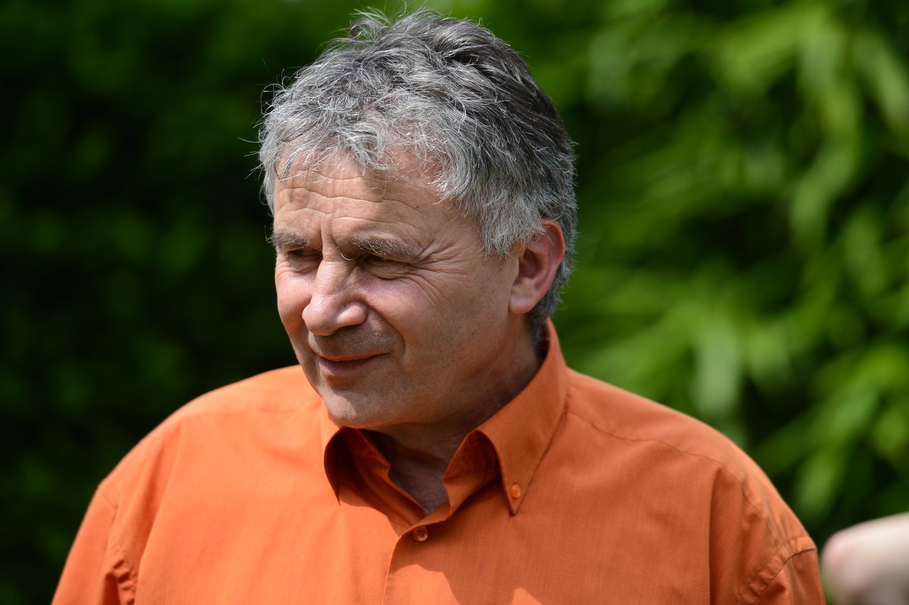 A man in an orange shirt smiles while standing outdoors with green foliage in the background.