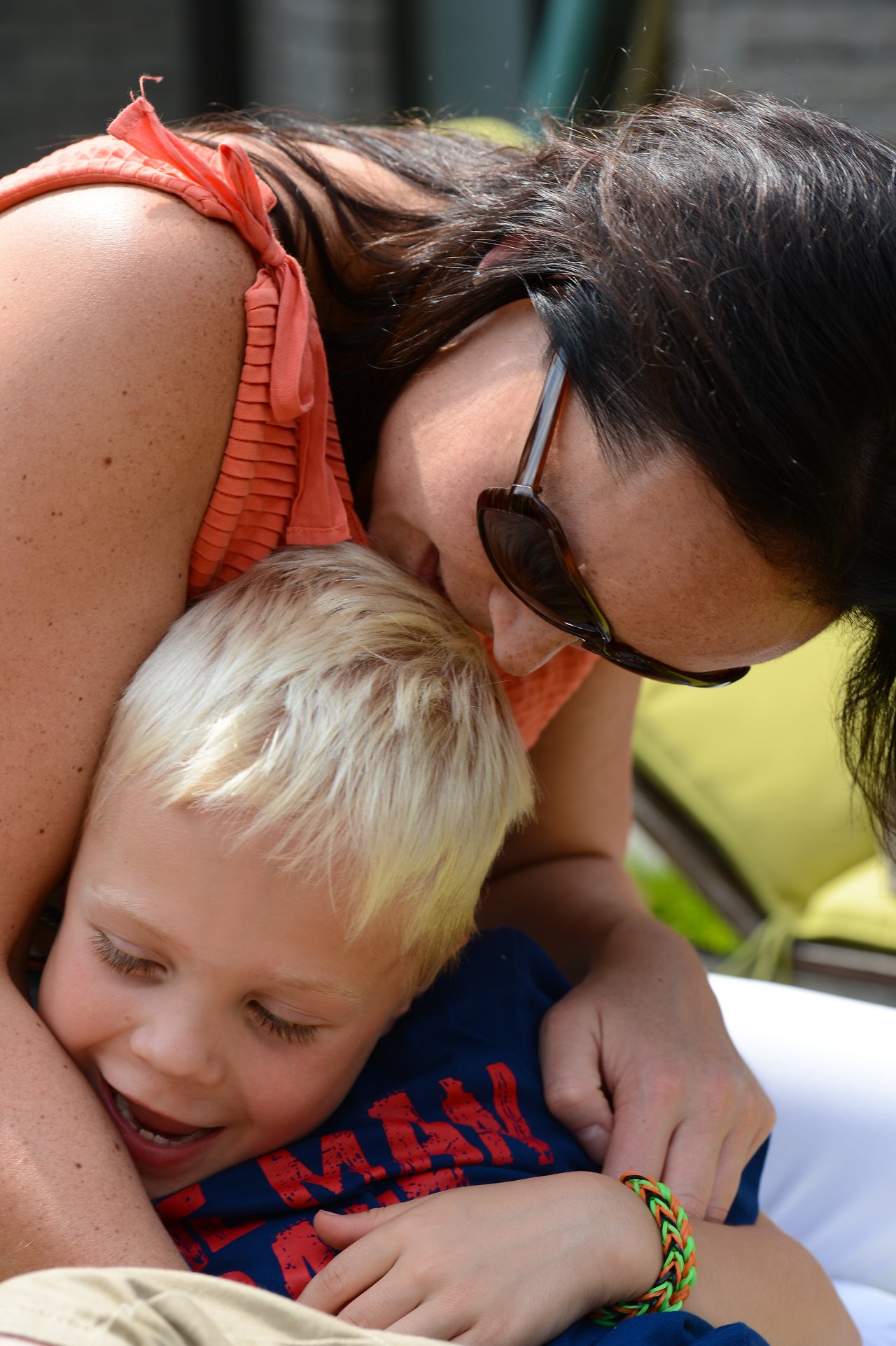A woman in sunglasses hugs a smiling young boy, who is wearing a blue shirt and colorful bracelet.