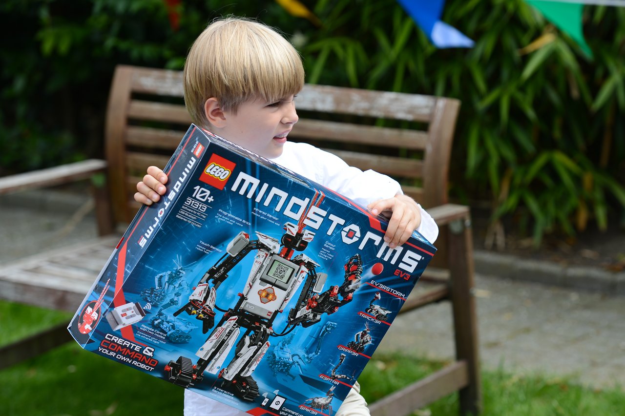 A child in a white shirt holds a large LEGO Mindstorms EV3 box while standing outdoors.