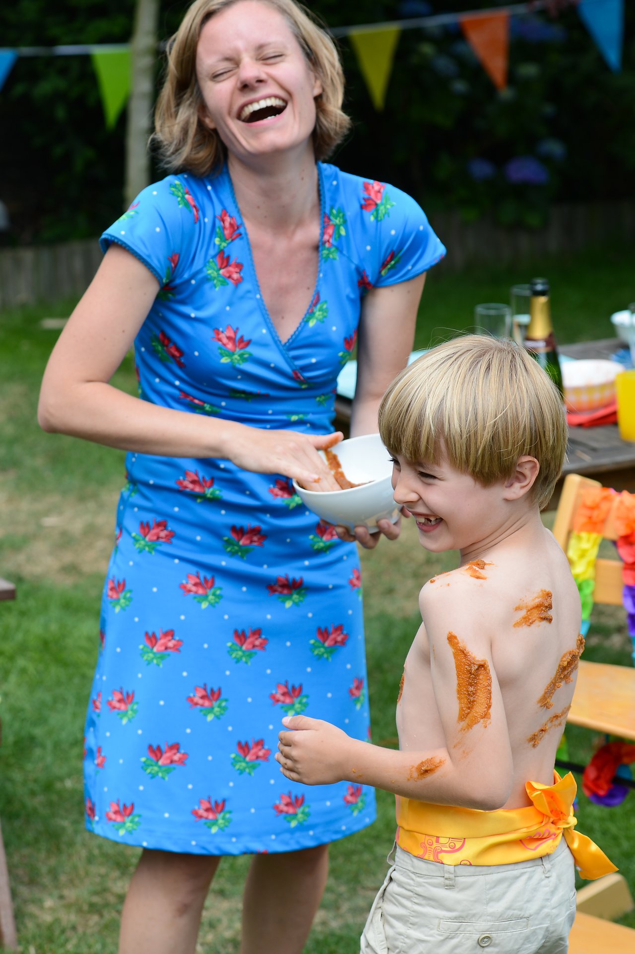 A woman laughs while applying sauce to a smiling child's bare back at an outdoor celebration.