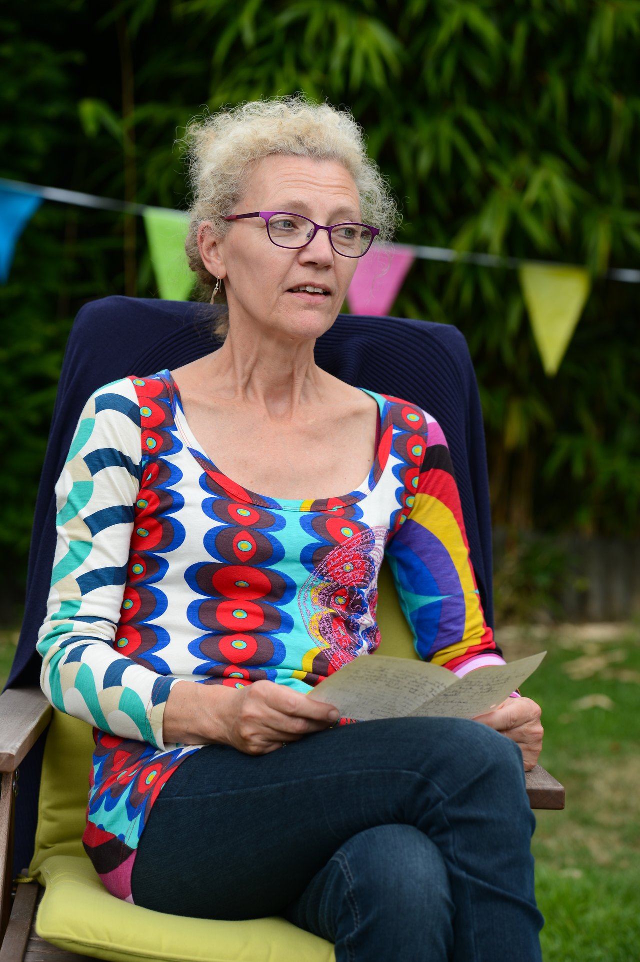 A woman wearing glasses and a colorful shirt sits outdoors, holding a piece of paper and speaking.