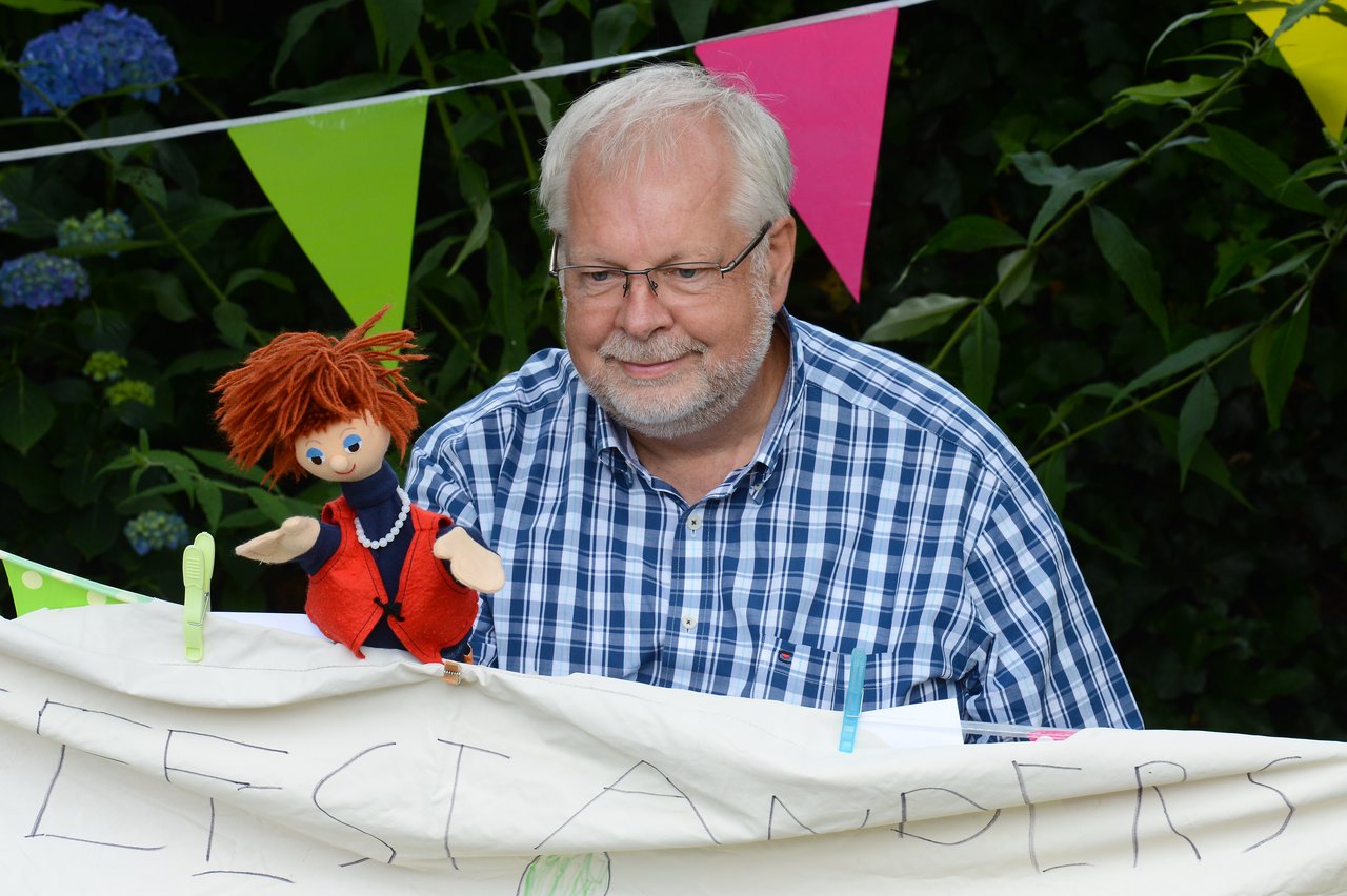 An older man in a plaid shirt holds a puppet behind a decorated banner at an outdoor celebration with colorful flags.