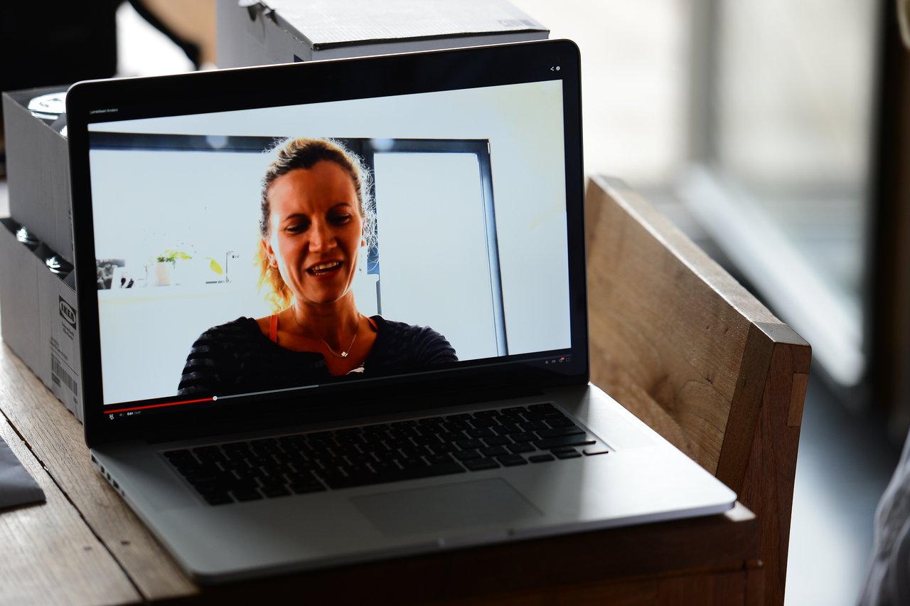 A laptop on a wooden table displays a video of a woman speaking in a well-lit room.