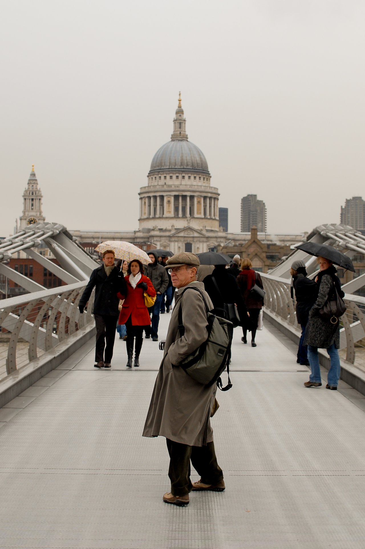 A man in a coat and backpack stands on a busy pedestrian bridge with people walking and carrying umbrellas.