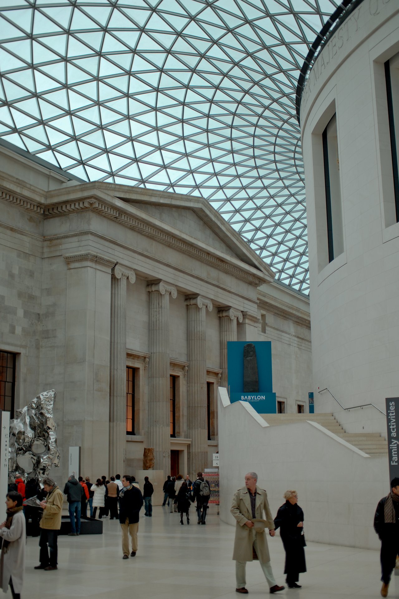 Visitors walk through the British Museum's Great Court, featuring a glass ceiling, classical architecture, and exhibition signs.