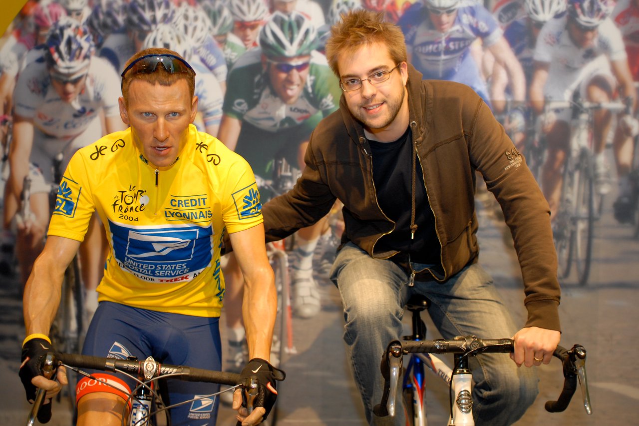 A man poses next to a lifelike Lance Armstrong figure on a bicycle, both appearing to ride in a race scene.