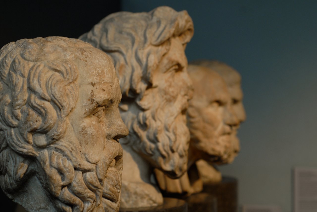 A row of ancient Greek marble busts with bearded faces, displayed in a museum setting.