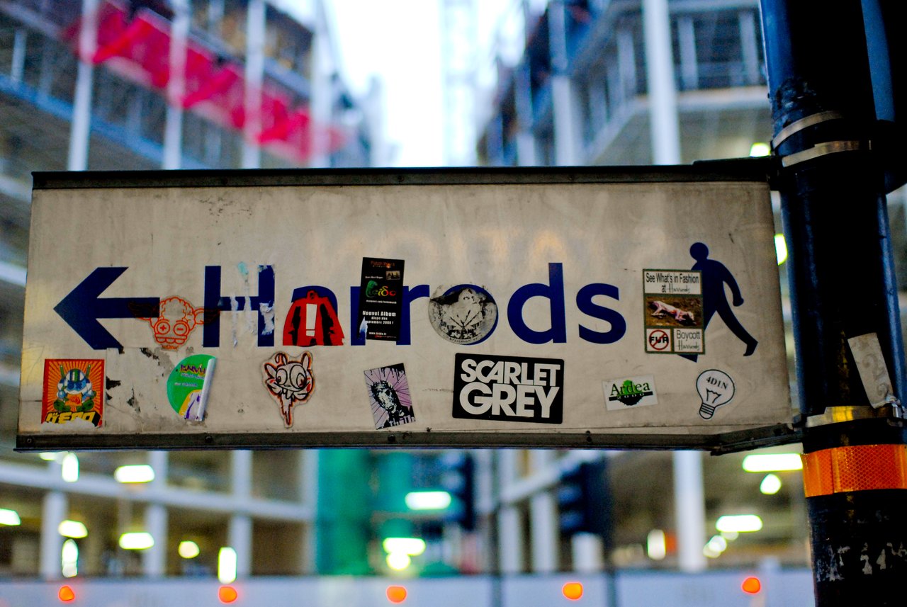 A street sign pointing to Harrods, covered in various stickers, with a blurred city background.