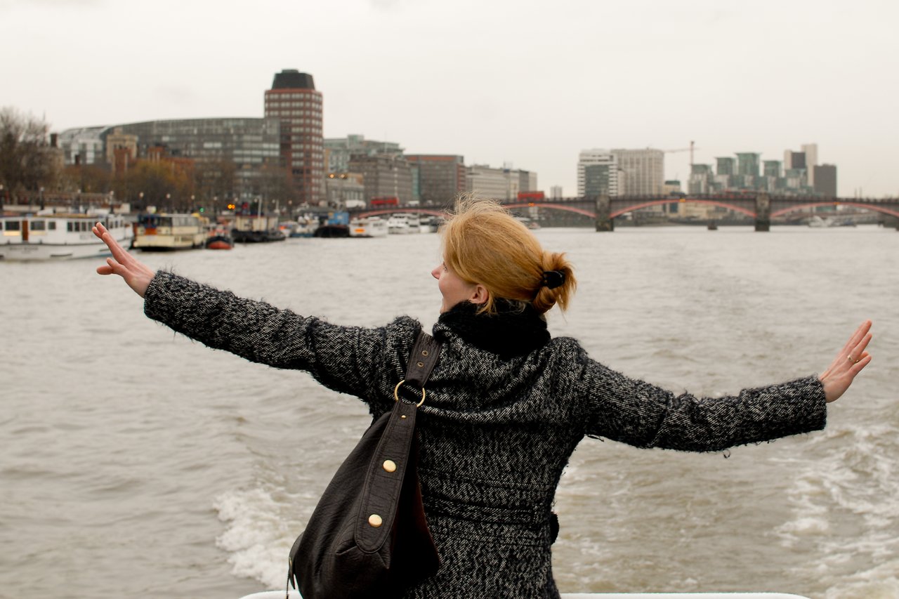 A woman with arms outstretched stands on a boat, facing the Thames River with a cityscape in the background.