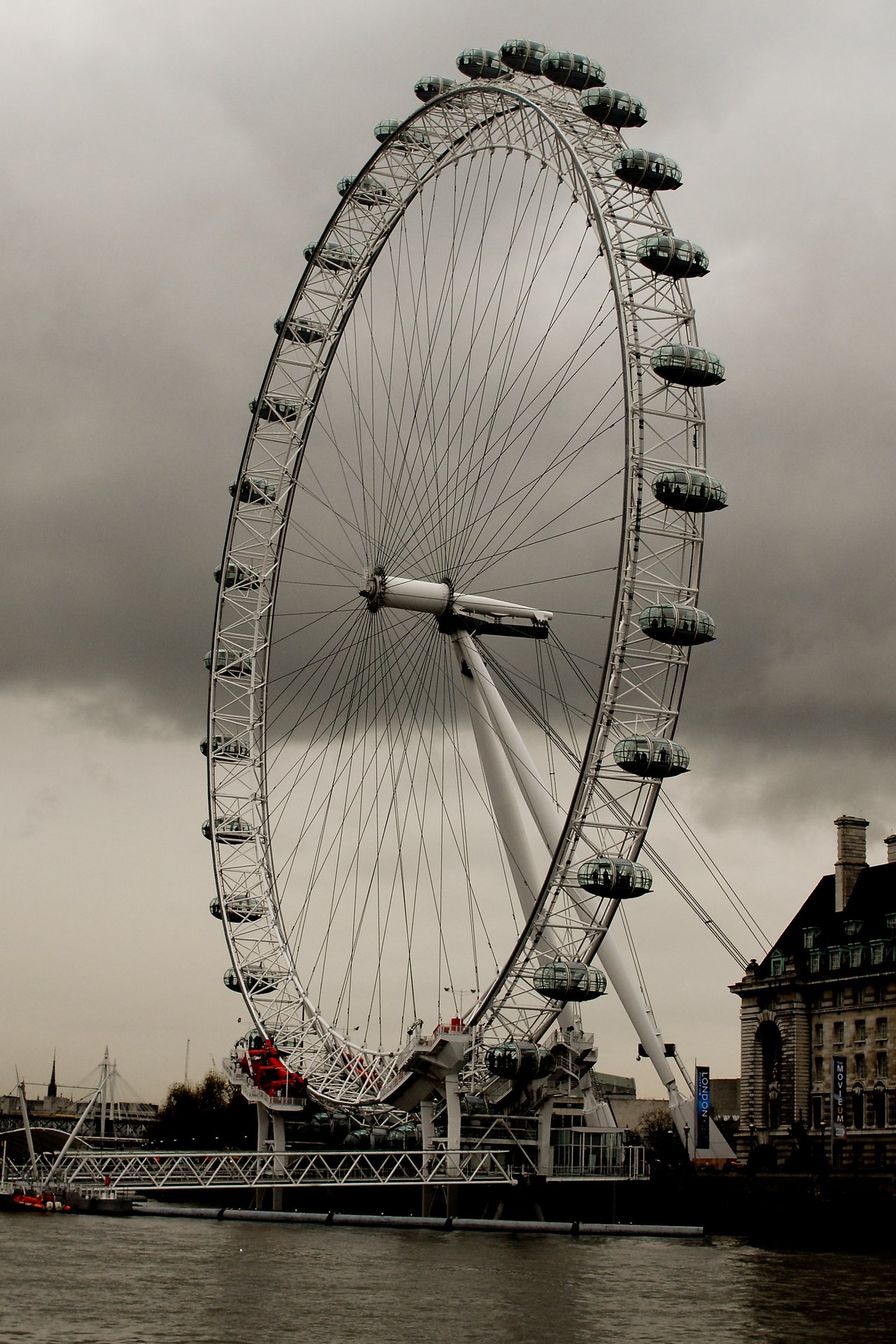 The London Eye, a large observation wheel with glass capsules, stands against a cloudy sky near the River Thames.