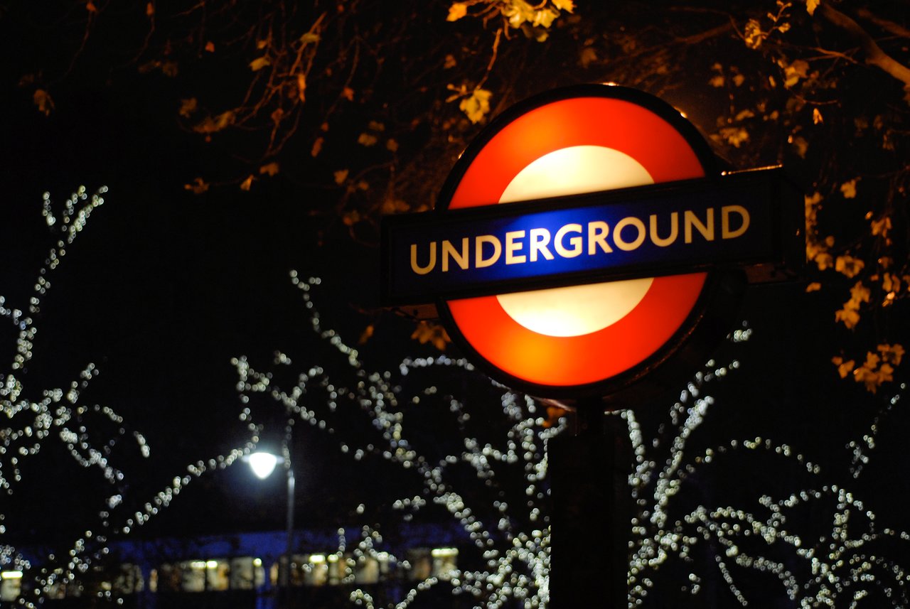 A London Underground sign glows at night, with trees and festive lights in the background.