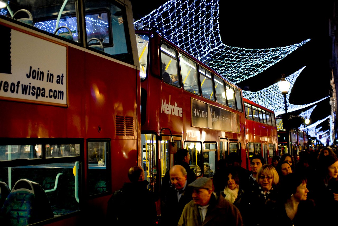 Busy crowd walking past red double-decker buses on Oxford Street at night, with festive lights overhead.
