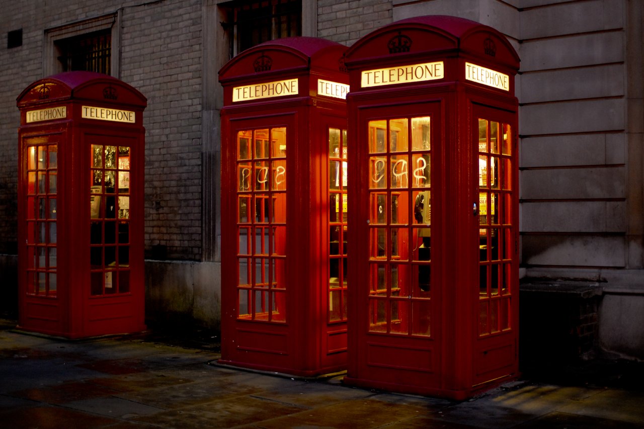 Three red British phone booths with glowing signs stand against a brick building at night.