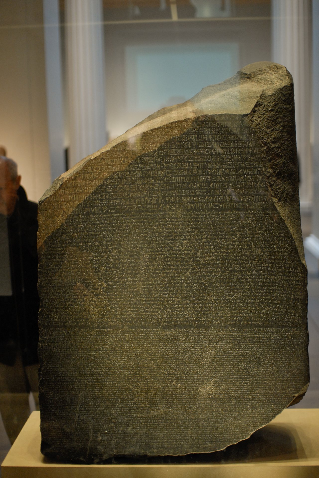 The Rosetta Stone, an ancient inscribed slab with three scripts, displayed in a museum with visitors in the background.