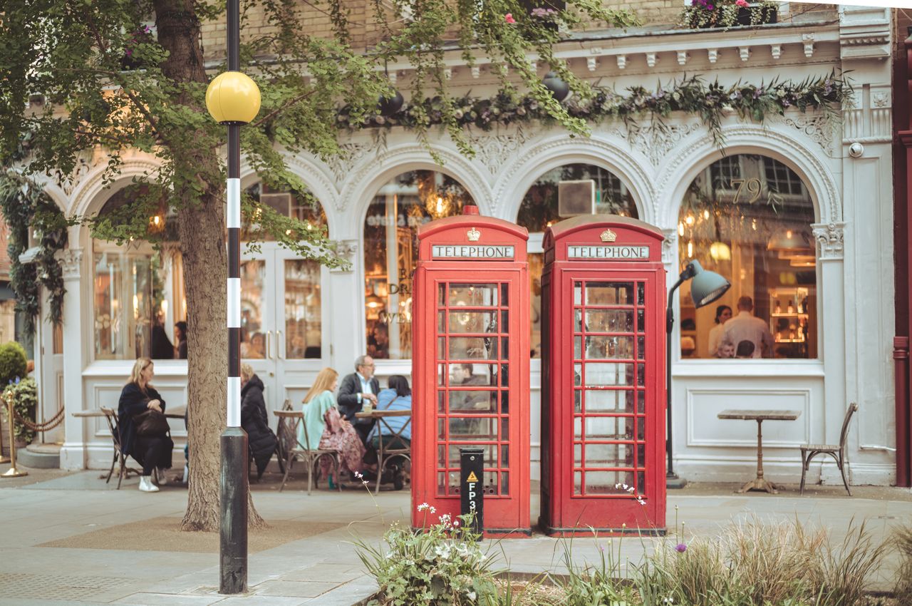 Two red phone booths with people sitting at outdoor restaurant tables in the background.