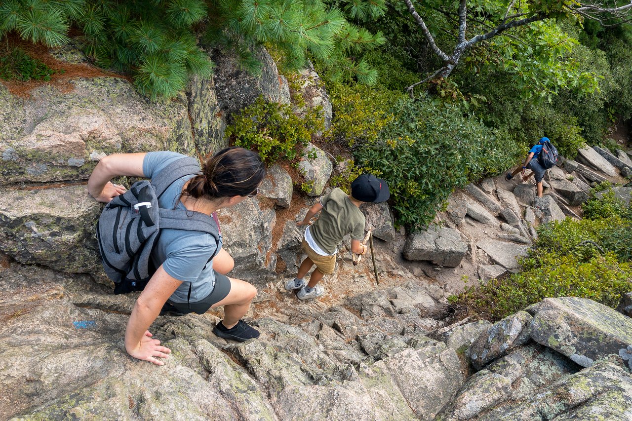 Three hikers carefully climb down a steep, rocky trail, using their hands and walking sticks for support.