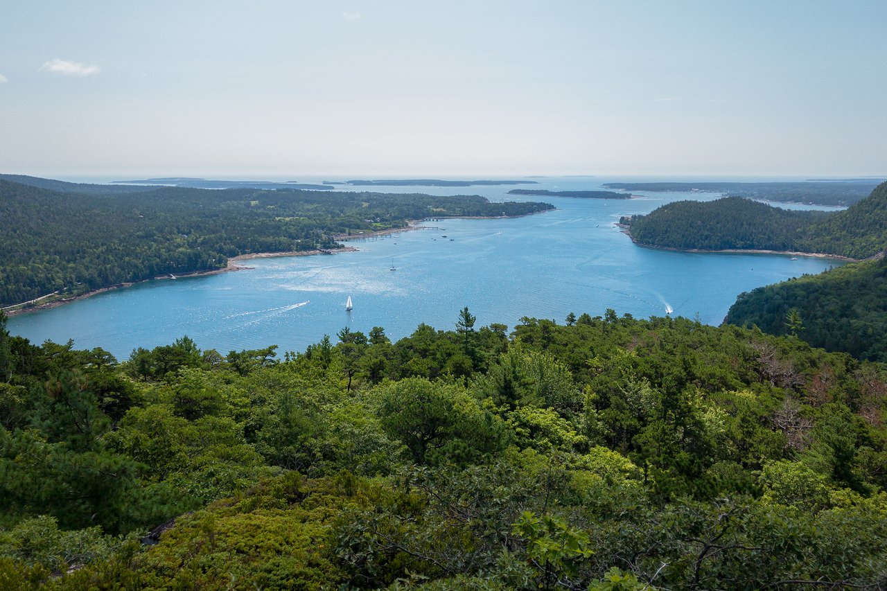 A sailboat moves across a wide blue bay, surrounded by green hills and distant islands under a clear sky.