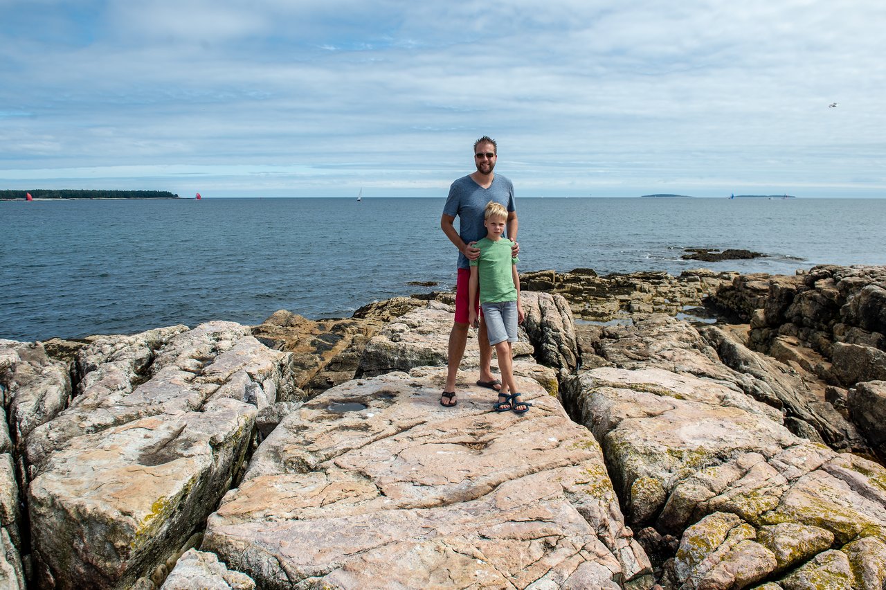 A man and a child stand on rocky terrain near the ocean, posing for a photo.