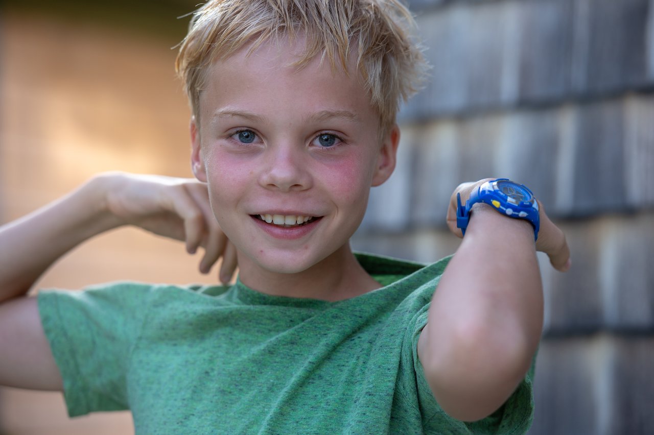 Smiling boy in a green shirt lifts his arms, wearing a blue watch on his wrist.