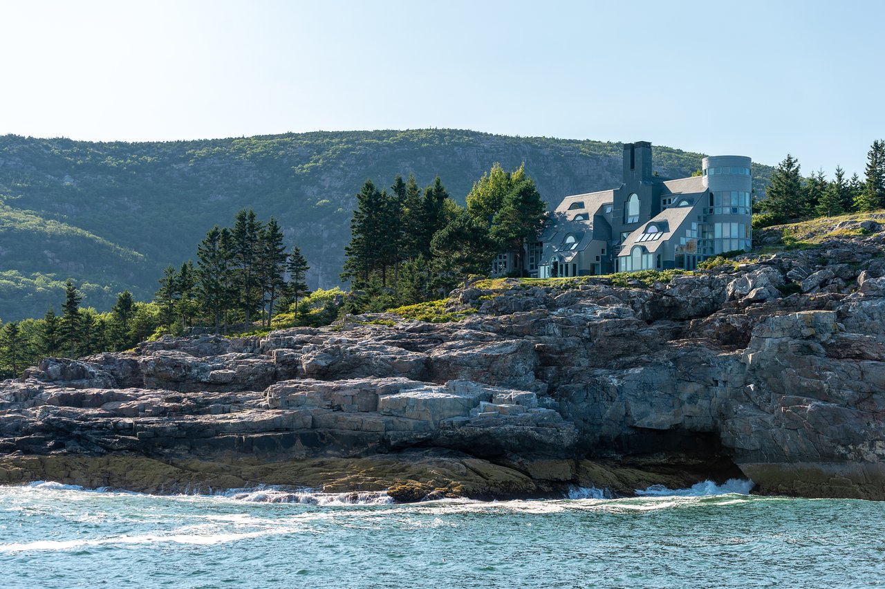 A large modern house sits on a rocky coastline, surrounded by trees, with mountains in the background.