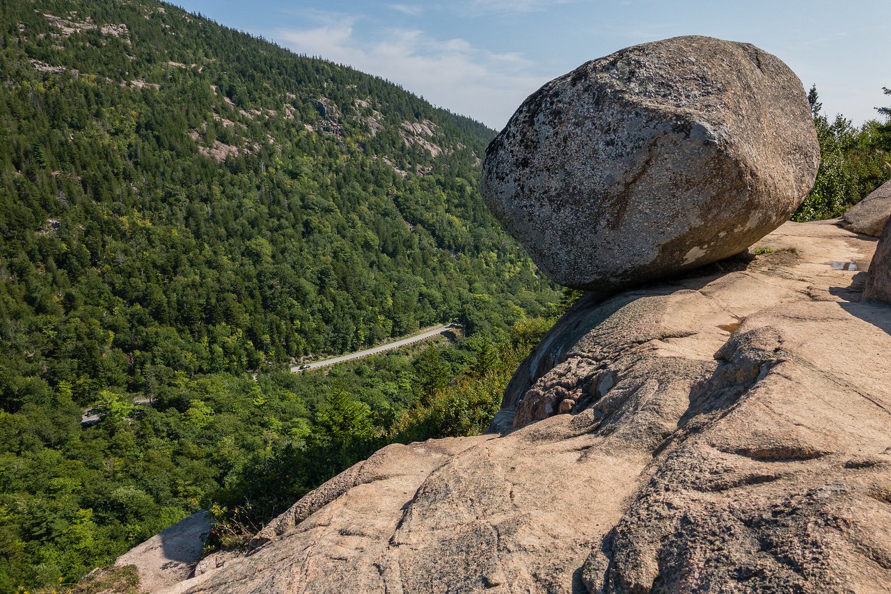 A large boulder rests on the edge of a rocky cliff overlooking a green forest and a winding road.