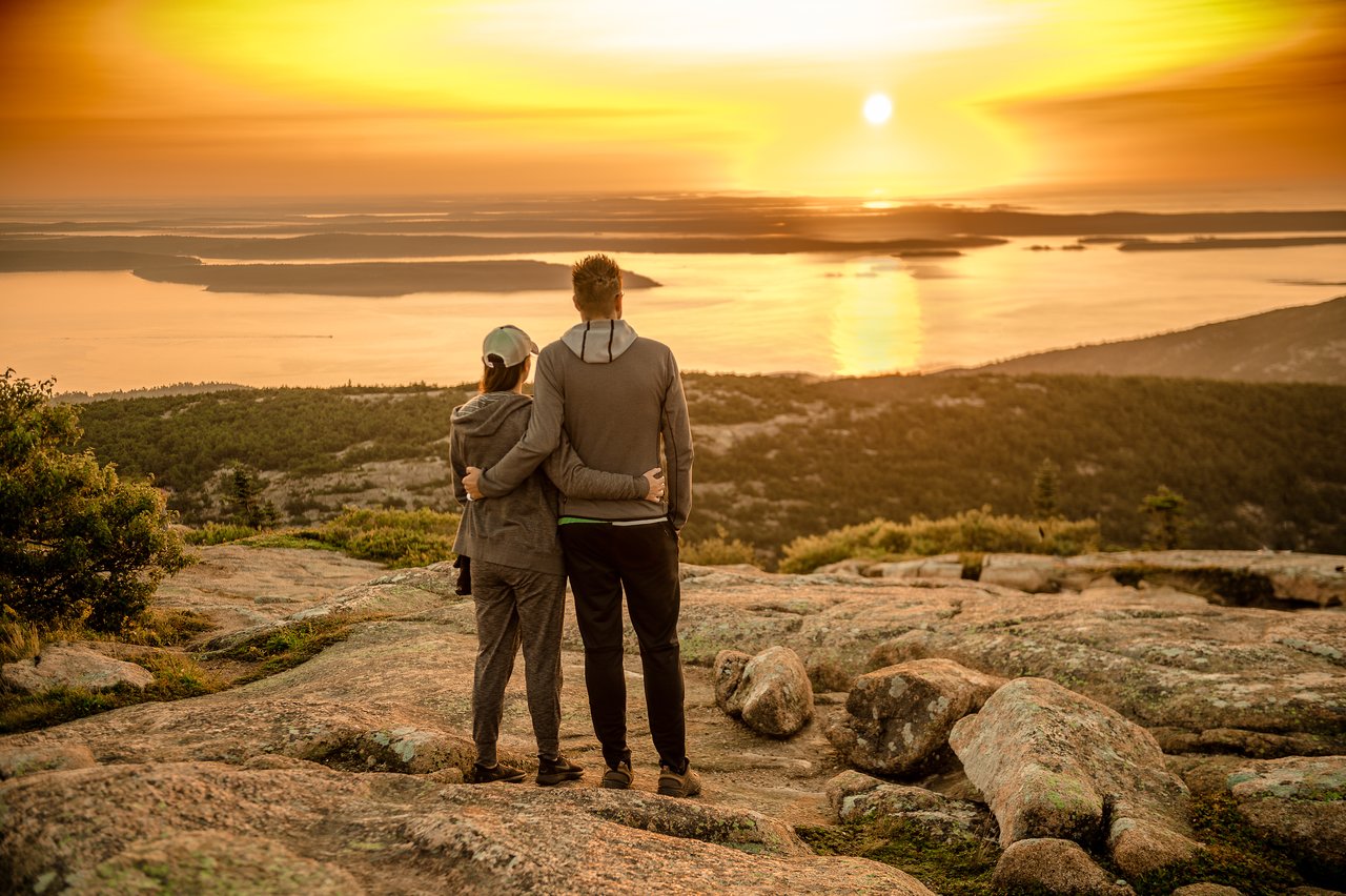A couple stands on a rocky hill, embracing as they watch the sunset over the water.