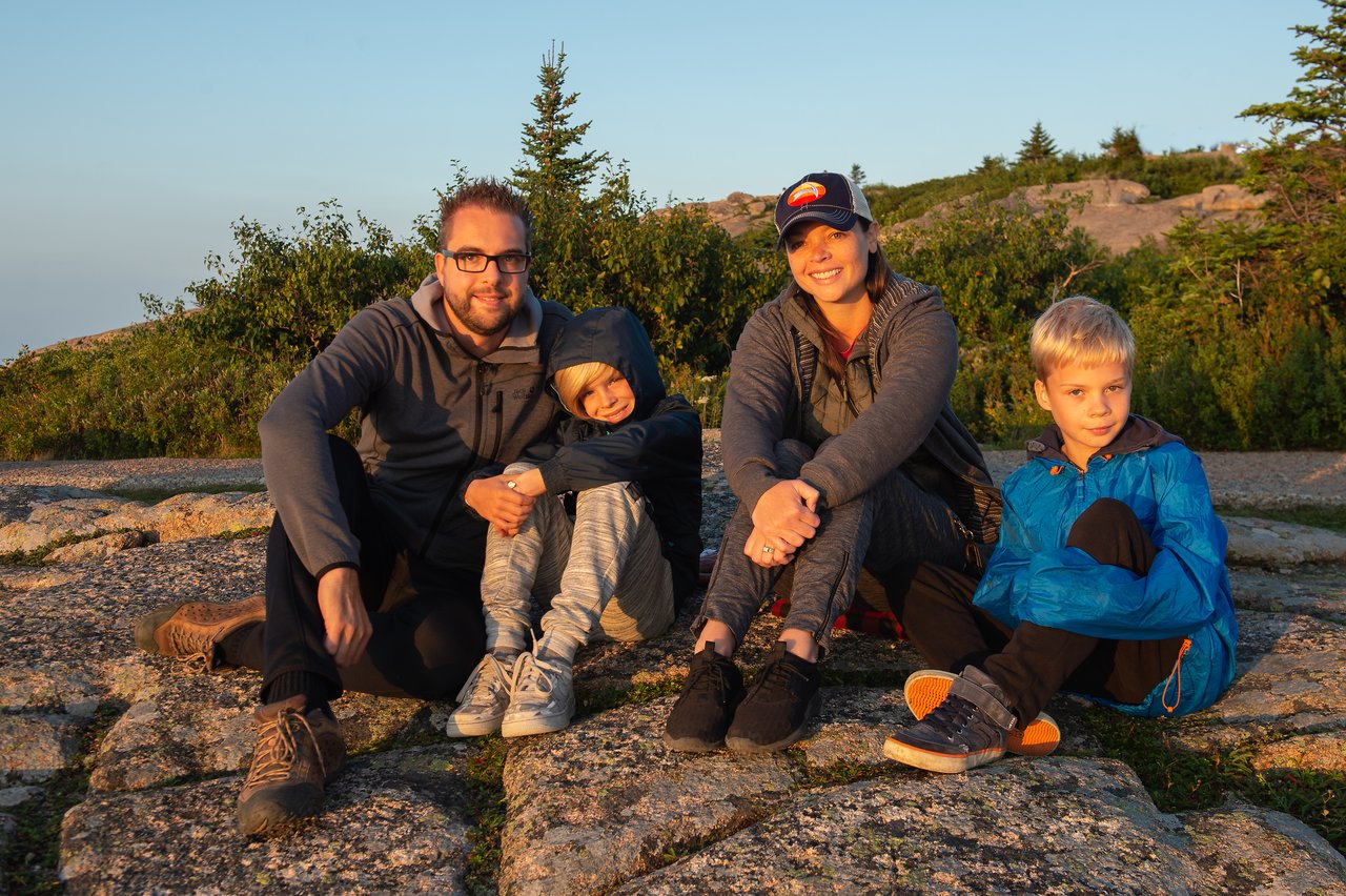 A family of four sits on rocky ground, dressed in jackets and hoodies, watching the sun rise.