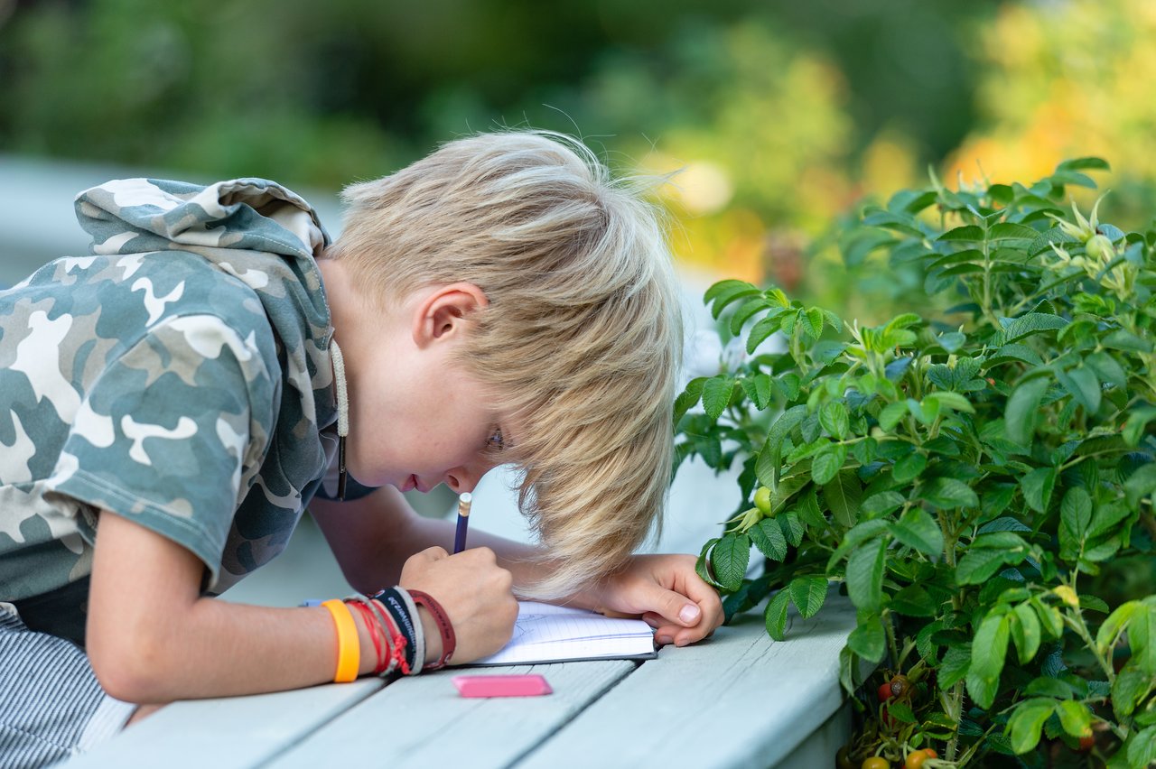 A young boy in a camouflage hoodie writes in a notebook with a pencil while sitting at a table.