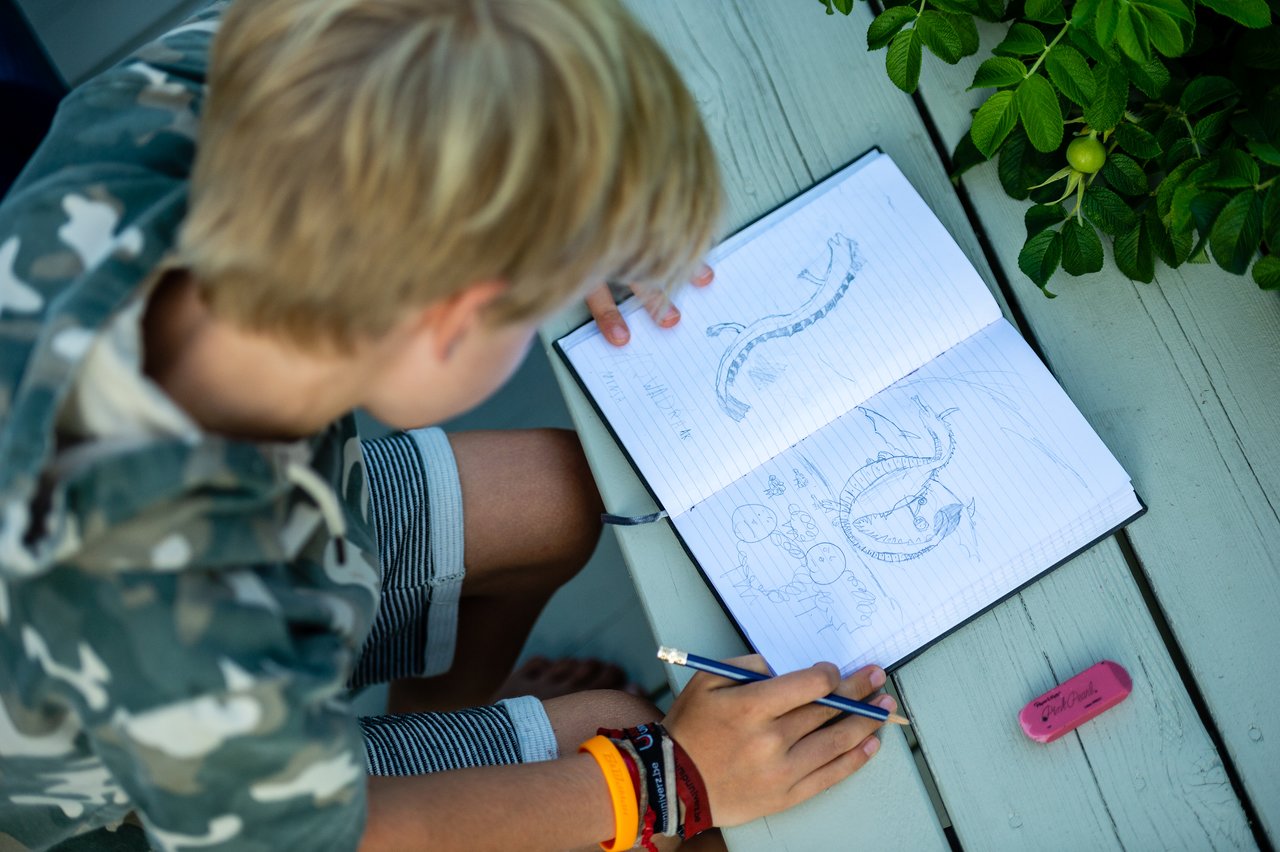 A child sits at a table, drawing detailed dragon sketches in a notebook with a pencil.