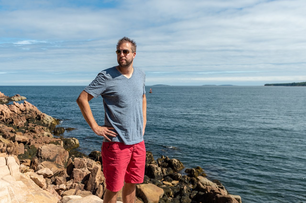 A man wearing sunglasses stands on rocky terrain near the ocean, looking into the distance with his hands on his hip.