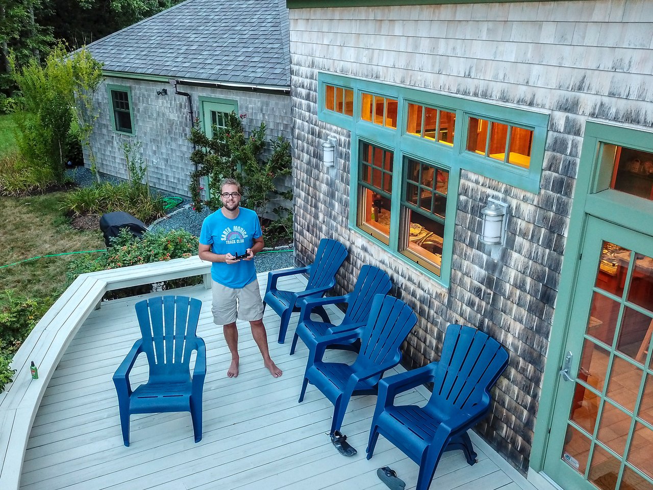 A man in a blue shirt stands on a deck holding a drone controller, looking up at the drone.