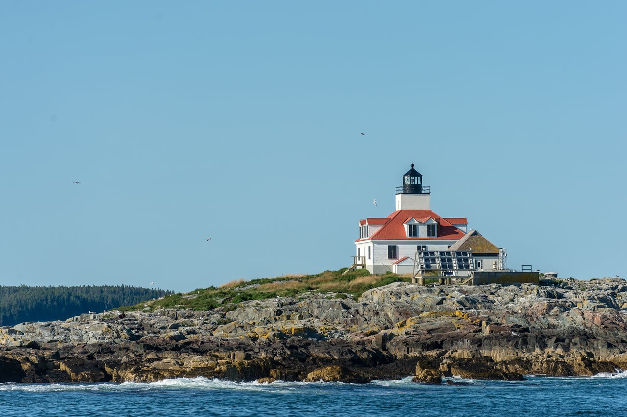 A white lighthouse with a red roof stands on a rocky coastline, surrounded by blue sky and ocean waves.