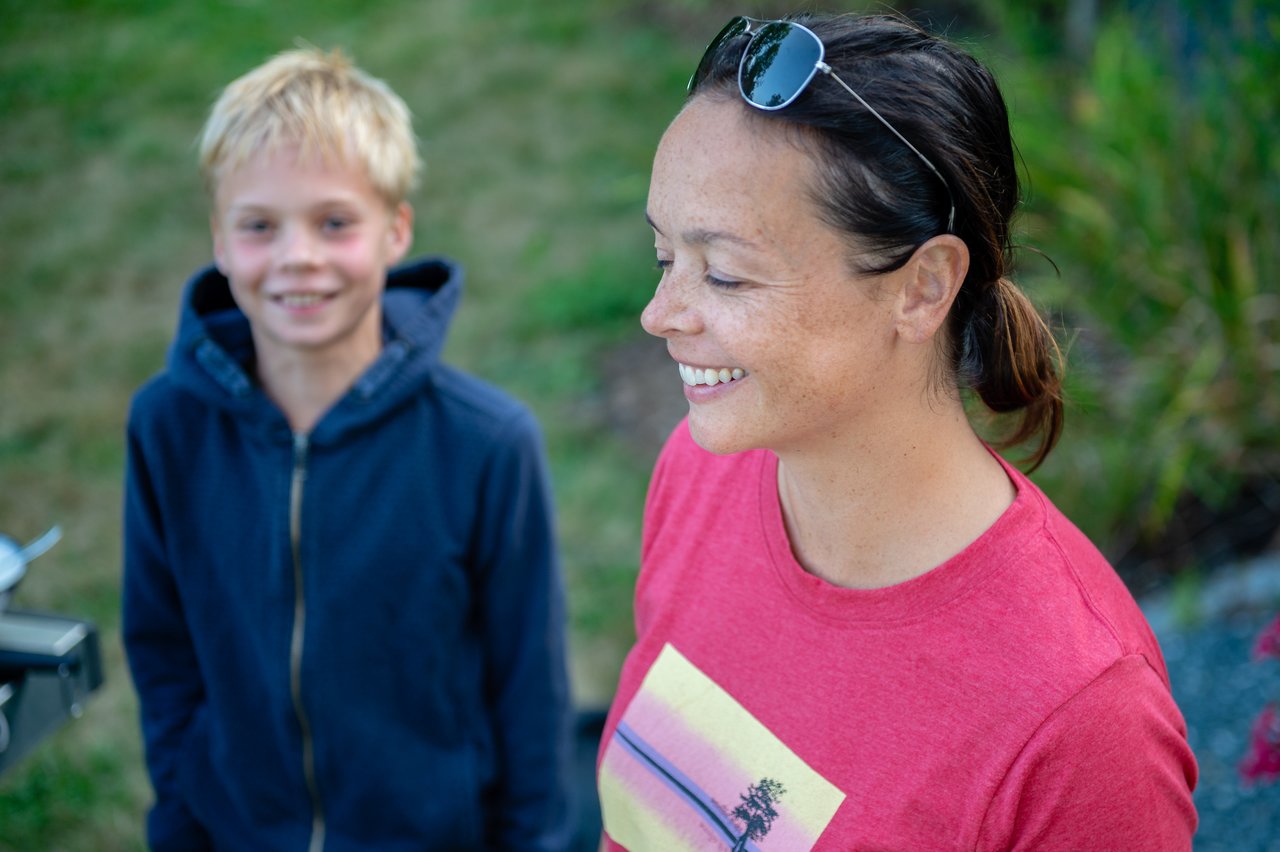 A smiling woman in a red shirt stands outdoors, with a young boy in a hoodie behind her.