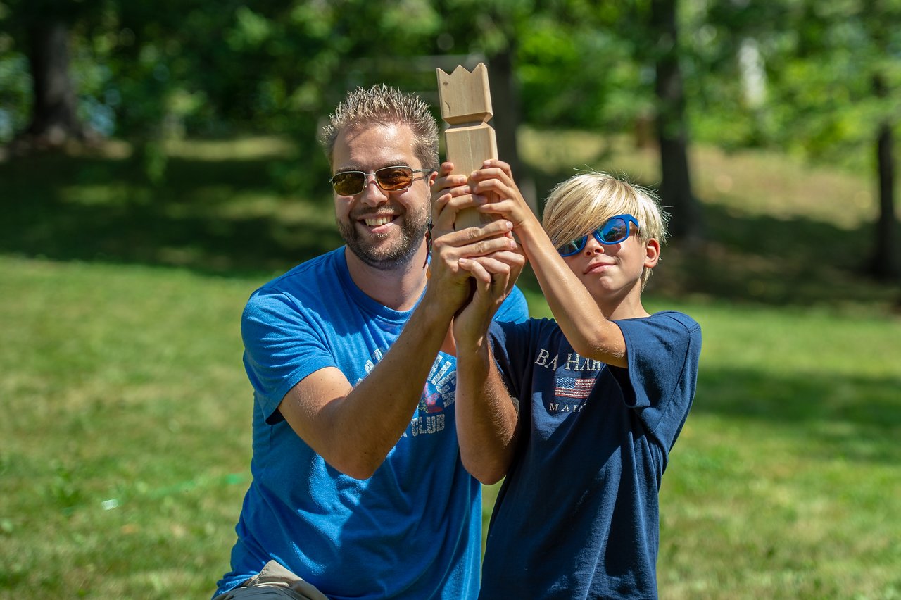 A man and a boy smile while holding up a wooden trophy together outdoors.