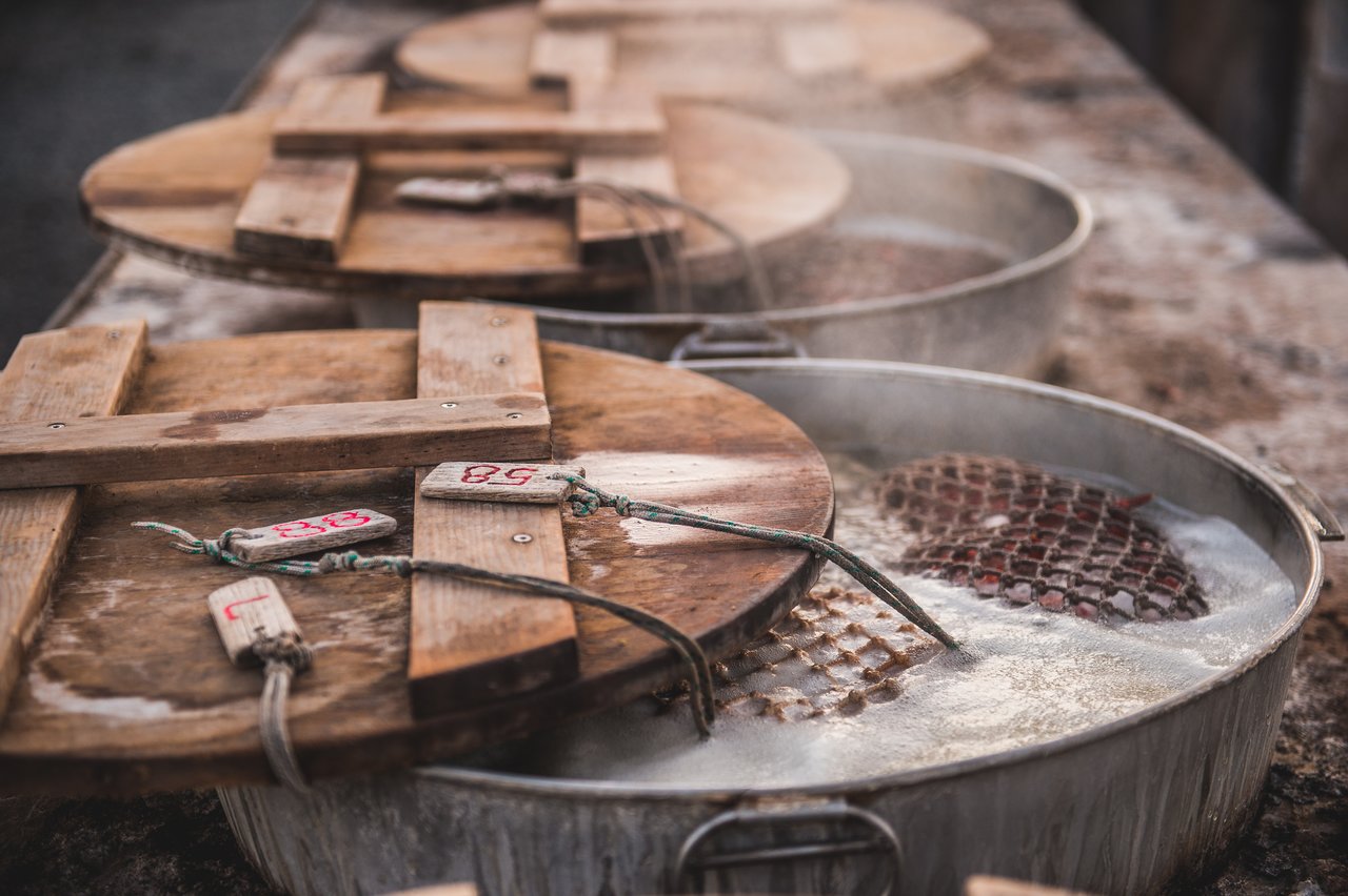 Large metal pots with wooden lids are boiling, with steam rising and numbered tags attached to the lids.