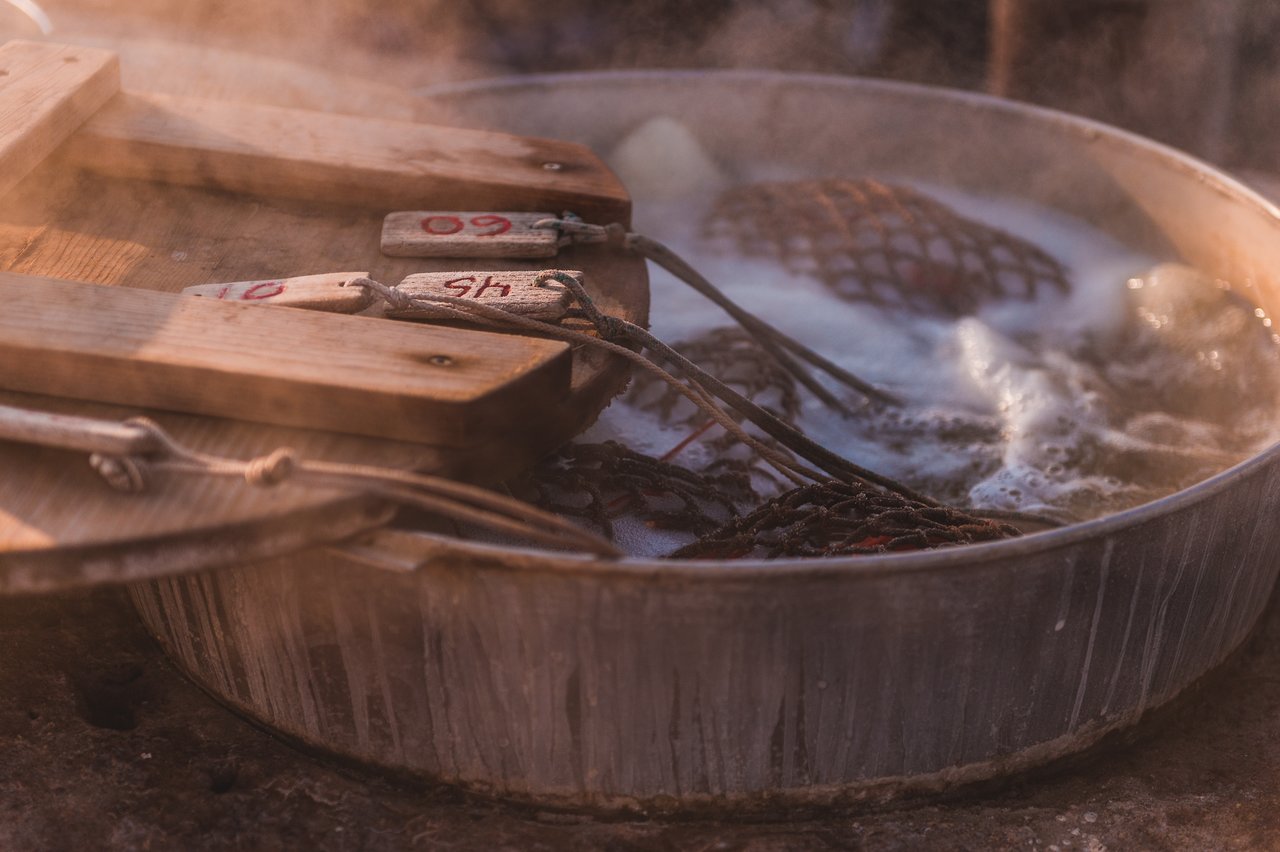 A large metal pot filled with boiling water contains netted bags, with wooden paddles resting on top.
