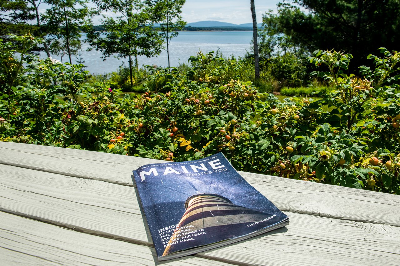 A travel magazine about Maine rests on a wooden table with greenery and a lake in the background.