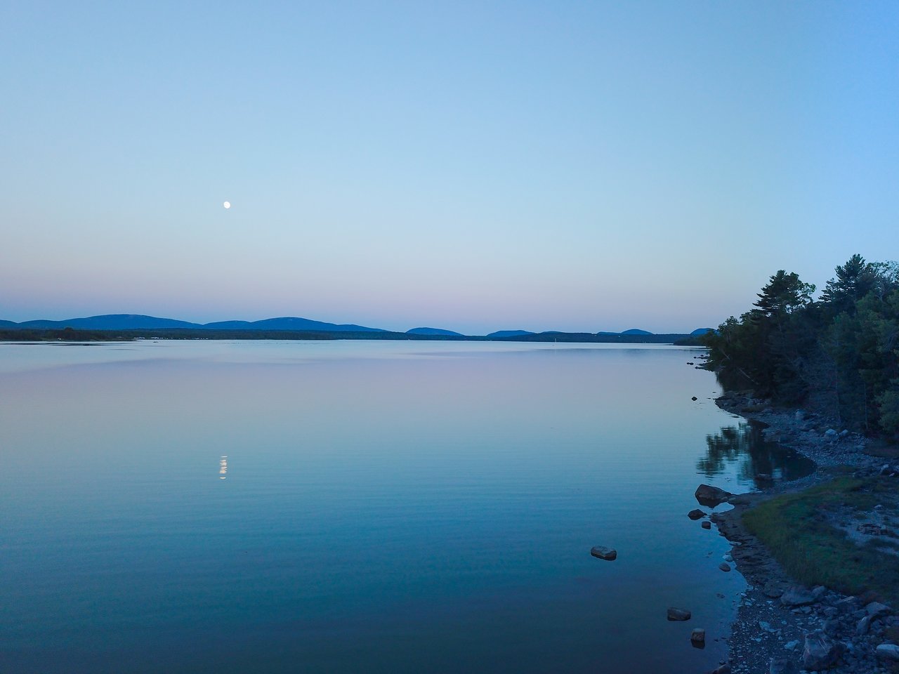 A calm lake reflects the moon, with a rocky shoreline and distant hills in the background.