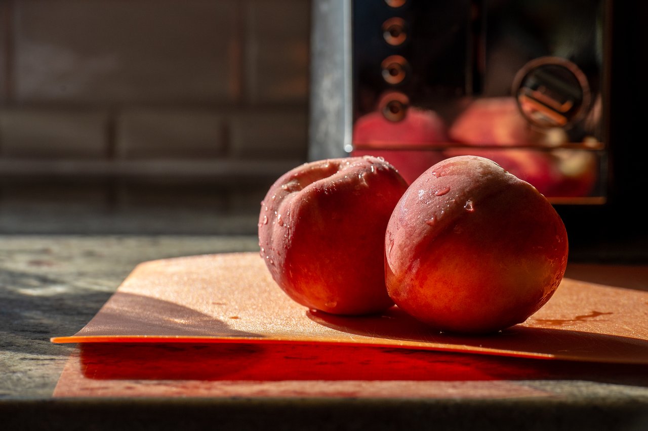 Two fresh peaches with water droplets rest on an orange cutting board on a kitchen counter.