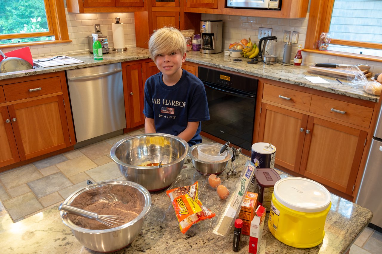 A child sits on a kitchen counter, surrounded by baking ingredients and mixing bowls, preparing to bake.