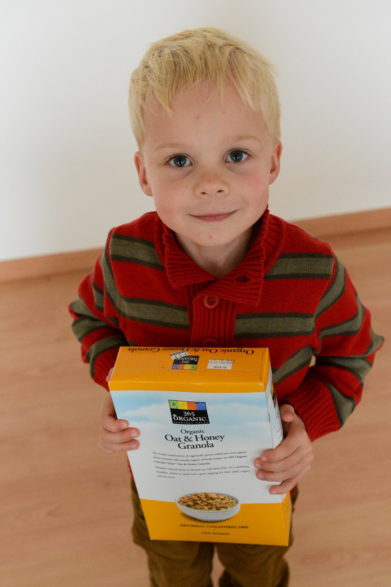 A young child in a striped sweater holds a box of oat and honey granola while looking at the camera.
