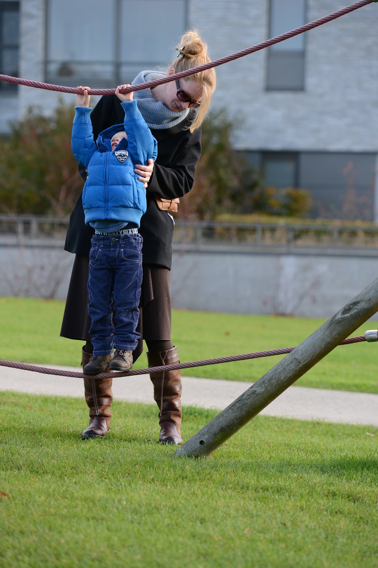 A woman helps a child balance on a rope structure at a playground, holding them for support.
