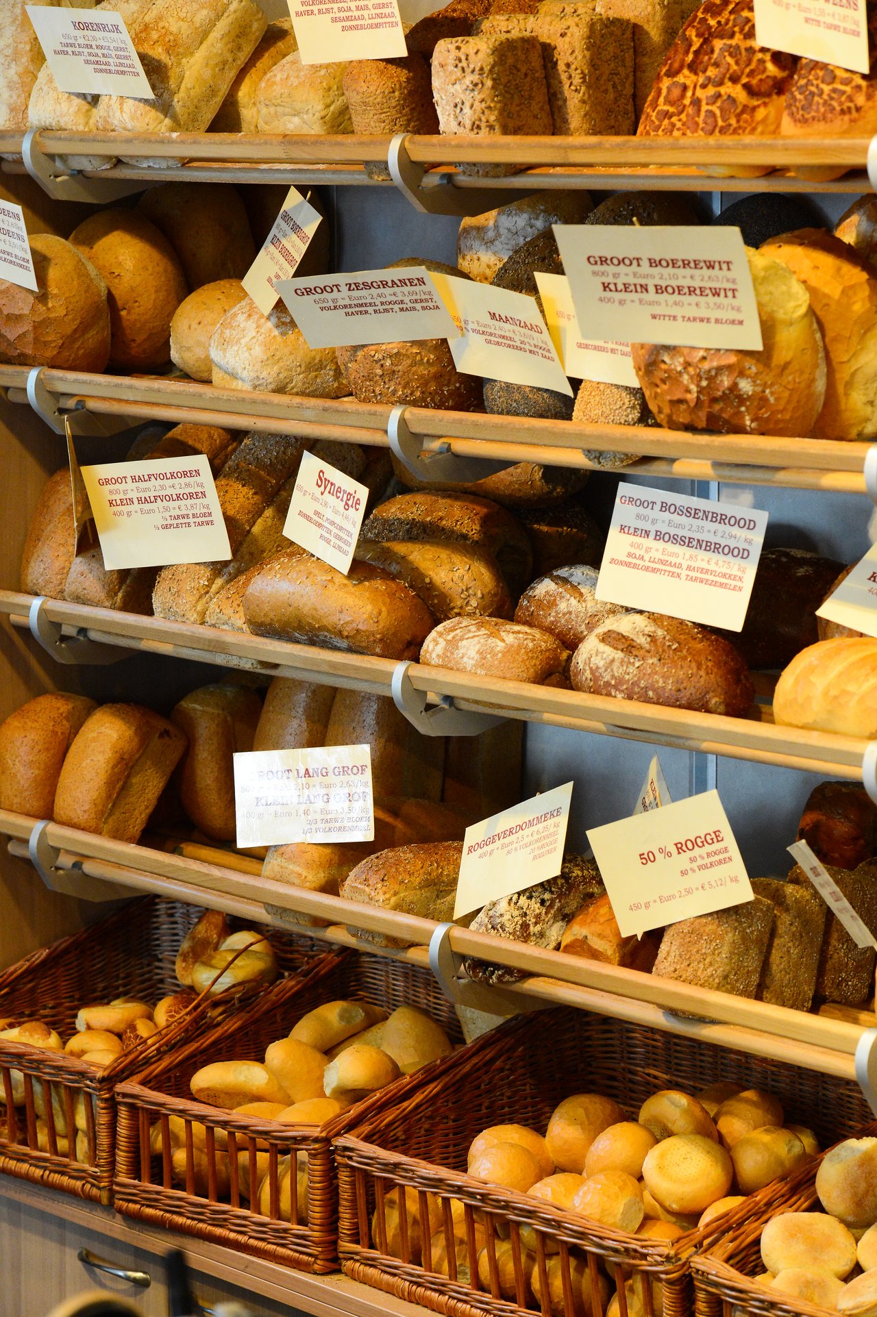 Shelves filled with various types of bread, including loaves and rolls, neatly arranged with labeled signs in a bakery.