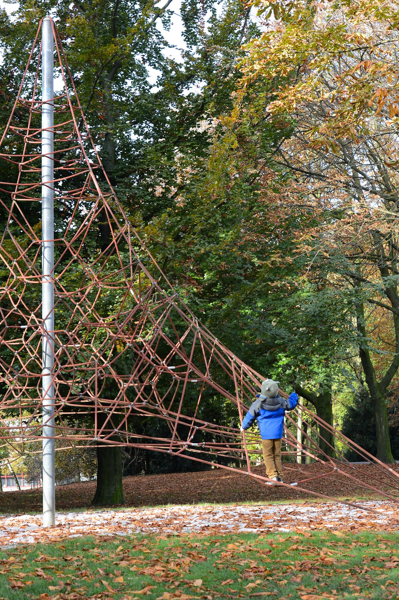 A child in a blue jacket climbs a rope playground structure in a park with autumn leaves on the ground.