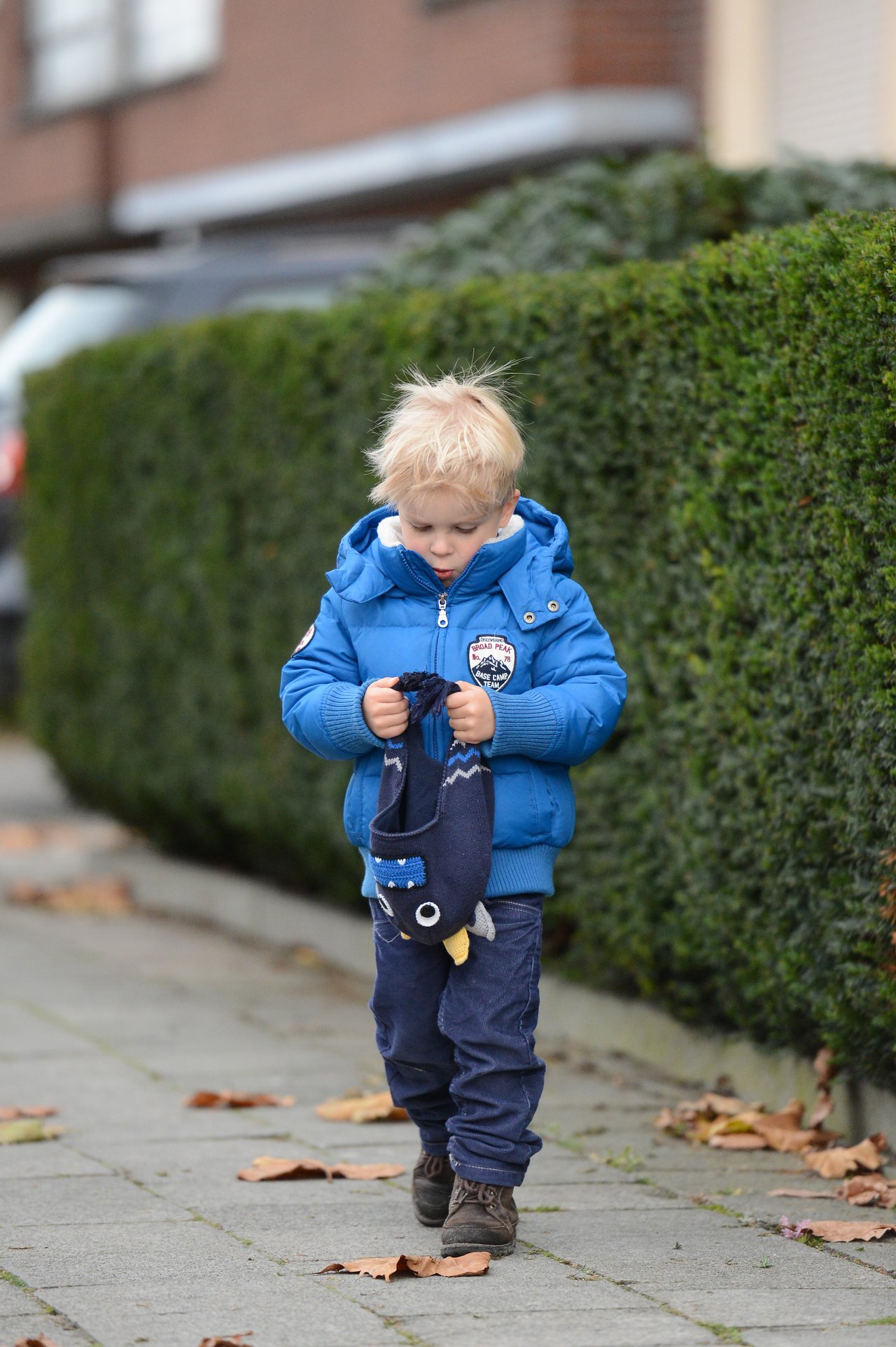 A young child in a blue jacket walks on a sidewalk, holding a hat with an animal design.