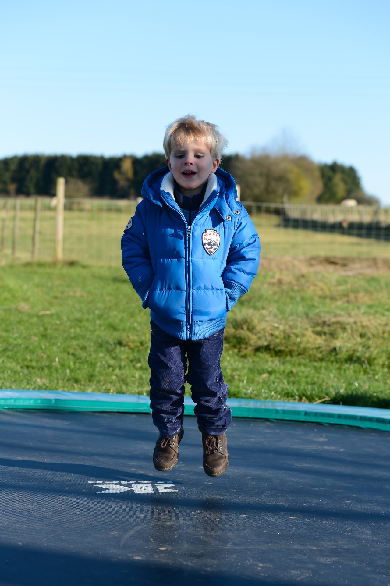 A young child in a blue jacket jumps on a trampoline outdoors during daytime.
