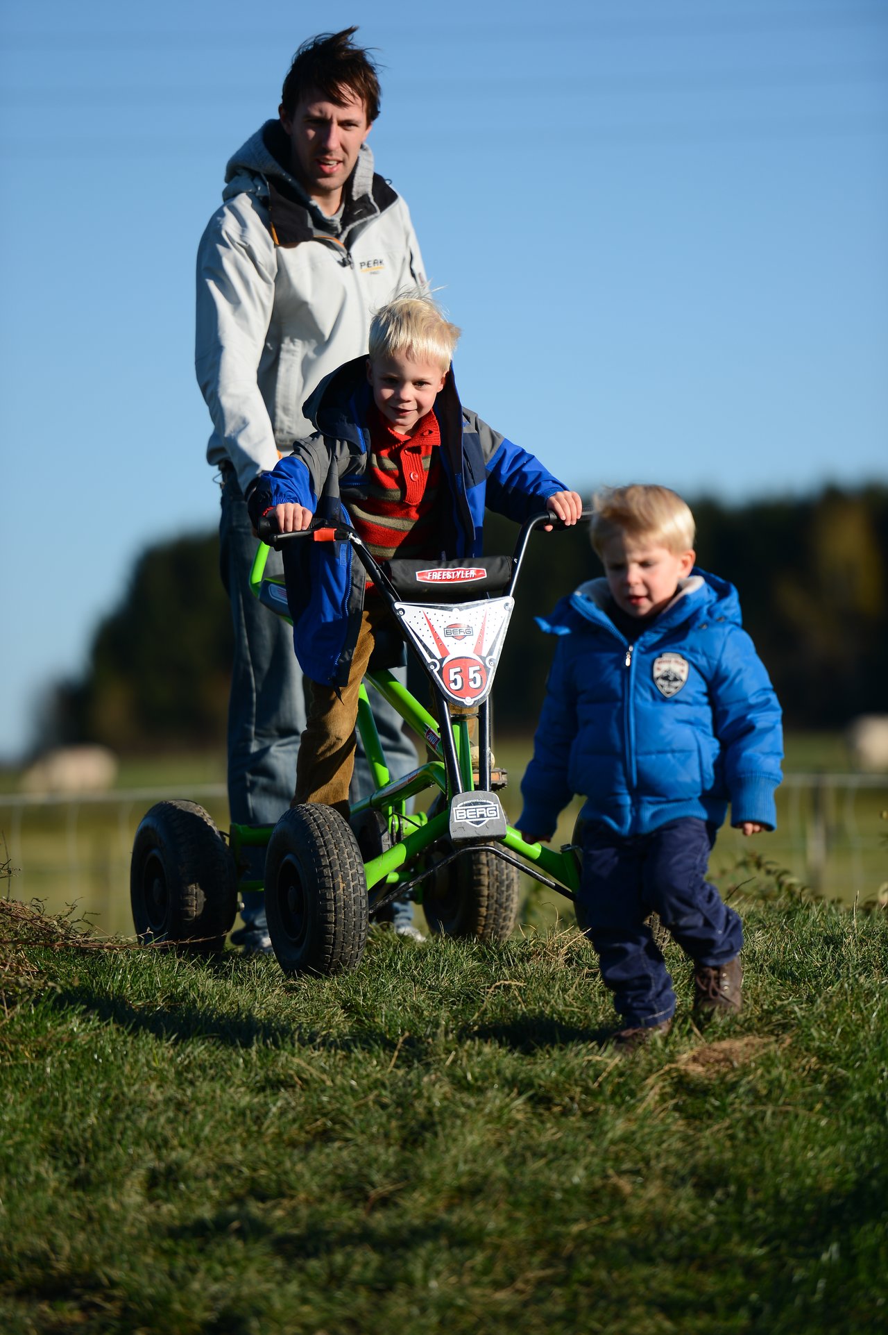 A man helps a child steer a green pedal kart while another child in a blue jacket runs ahead.