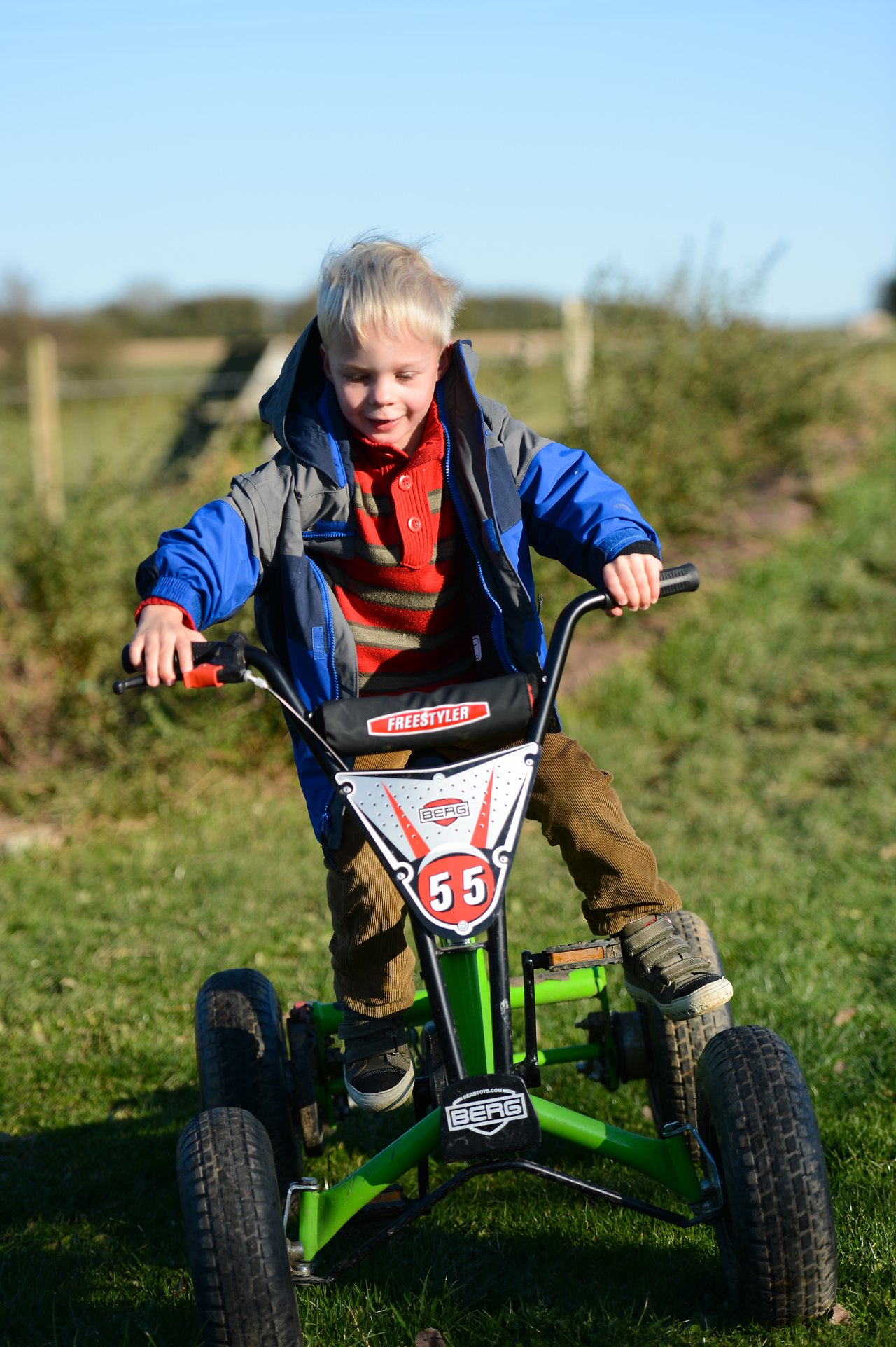 A young child rides a green pedal go-kart on a grassy path, gripping the handlebars and looking forward.
